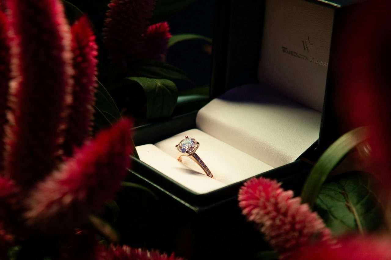 a side stone diamond engagement ring in a ring box surrounded by flowers