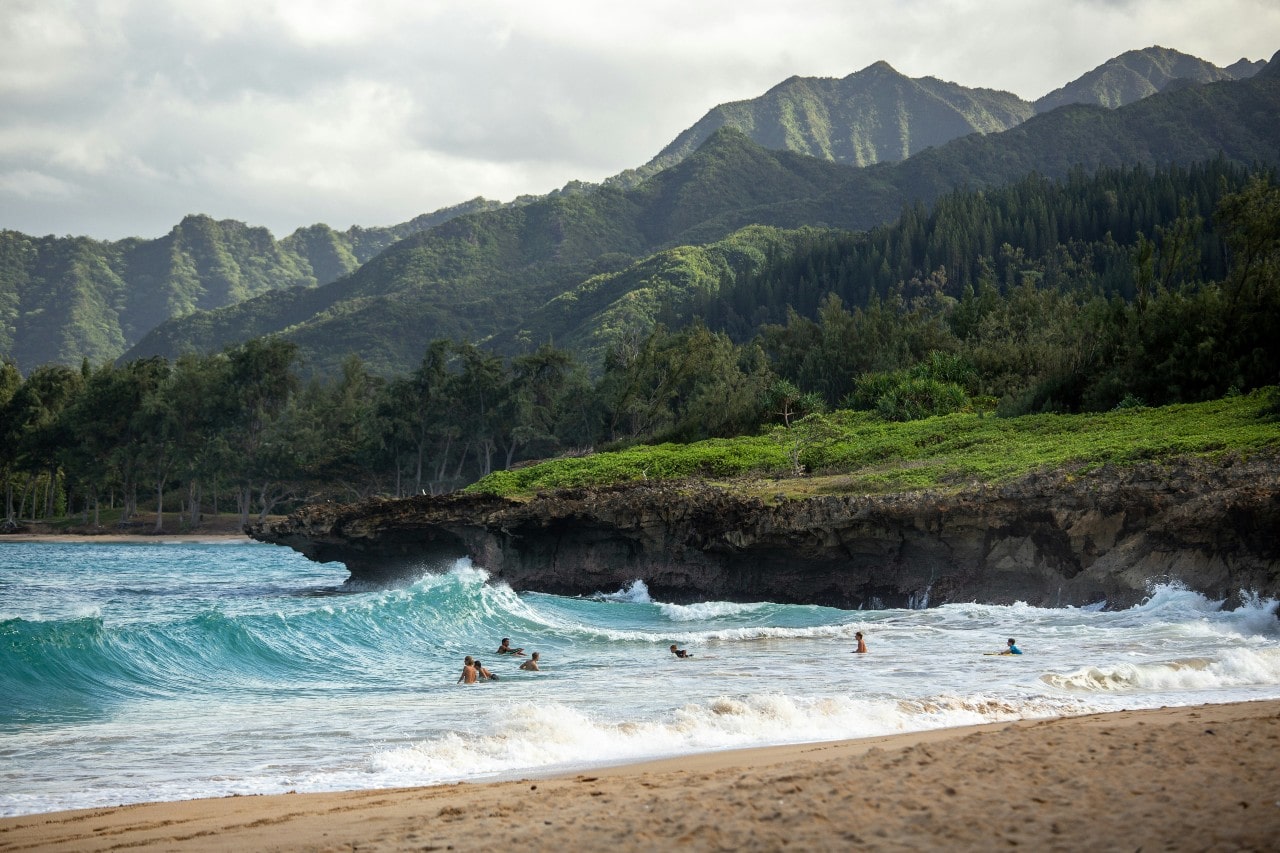 a beach in Hawaii with people swimming in the waves and mountains in the background