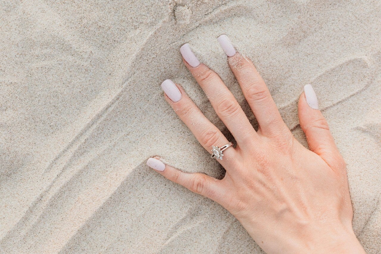 a woman’s hand in the sand wearing a white gold engagement ring a woman’s hand in the sand wearing a white gold engagement ring