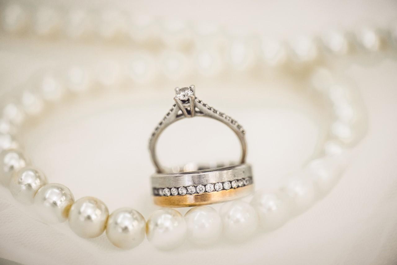 An engagement ring and a wedding band sit among a pearl necklace on a white cloth. An engagement ring and a wedding band sit among a pearl necklace on a white cloth.