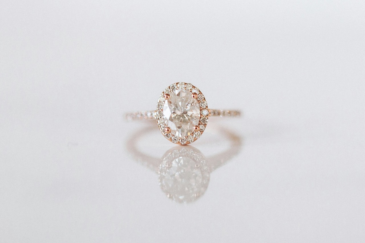 A rose gold halo engagement ring with an oval cut center stone displayed on a reflective white background. A rose gold halo engagement ring with an oval cut center stone displayed on a reflective white background.