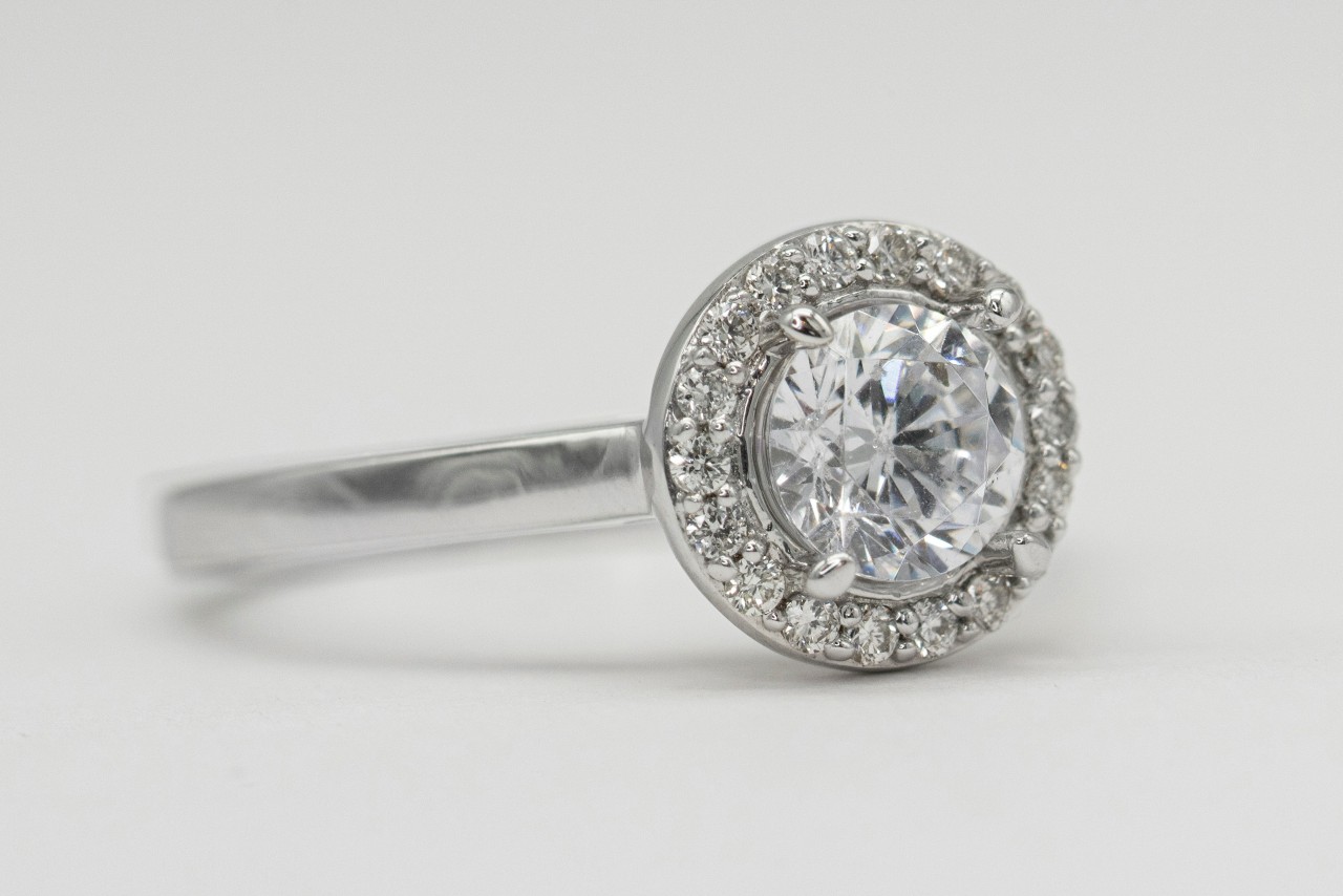 A close-up of a white gold bezel set halo engagement ring on a white surface. A close-up of a white gold bezel set halo engagement ring on a white surface.